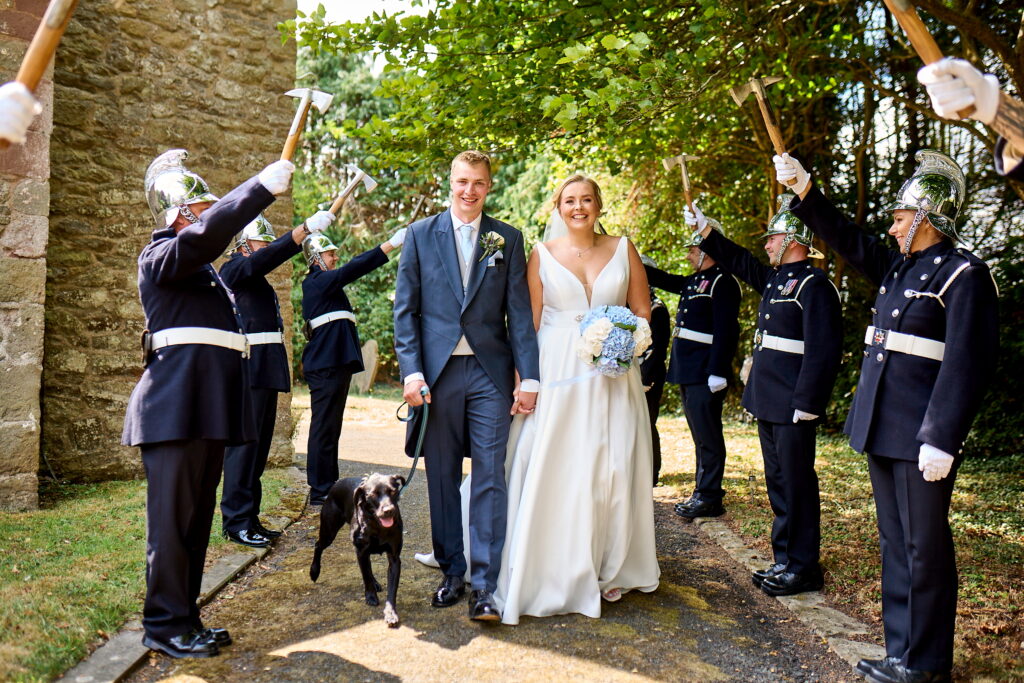 Bride in a white wedding gown and groom in a gray suit walk together as uniformed soldiers cheer with raised axes along a stone wall; a black dog accompanies them, bouquet of blue hydrangeas in the bride's other hand.