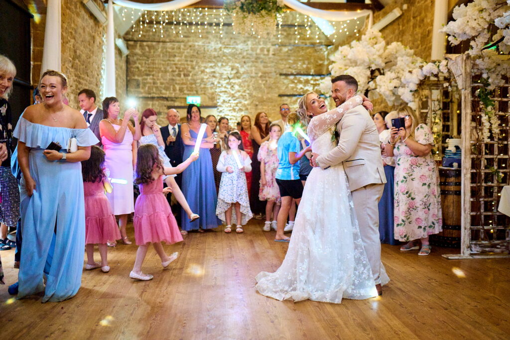 Bride and groom share a kiss during a dance on the wedding floor, surrounded by guests in colorful dresses under string lights.